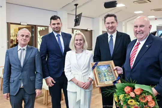 Grupa pięciu osób pozujących z obrazem i bukietem kwiatów.
