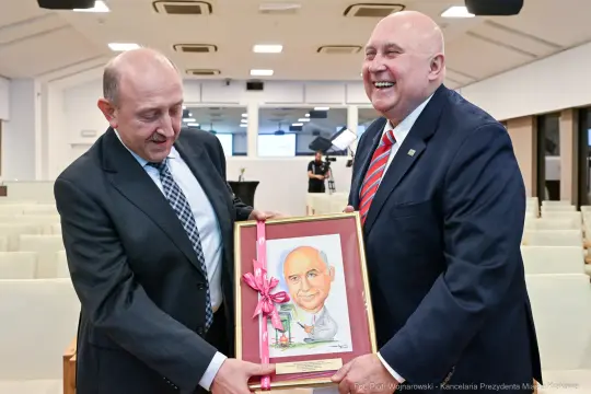 Dwaj mężczyźni wręczają sobie portret z okazji uroczystości. Obaj uśmiechnięci.