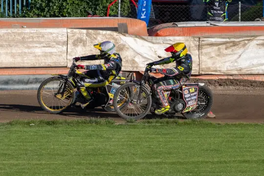 Dwaj zawodnicy na motocyklach ścigają się na torze żużlowym.