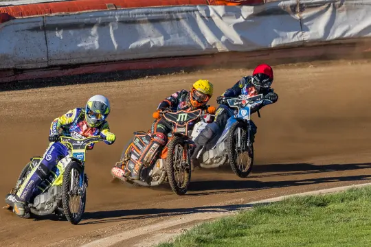 Trzech zawodników na motocyklach ściga się na torze żużlowym.