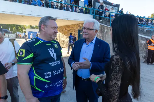 Mężczyźni rozmawiają w sportowych koszulkach, w tle tłum i stadion.