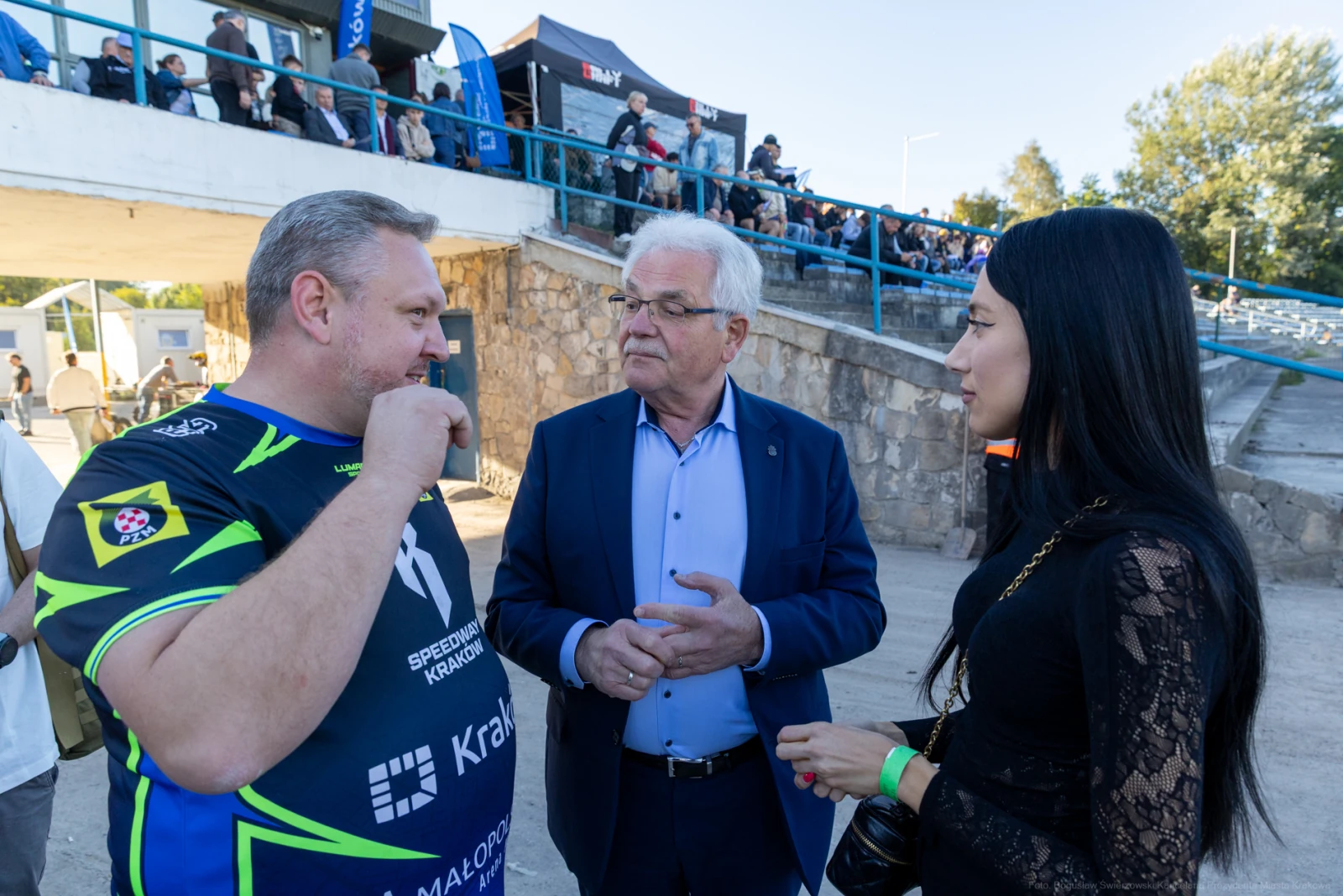 Trzech ludzi rozmawia na tle stadionu, widać kibiców na trybunach.  Autor: Bogusław Świerzowski