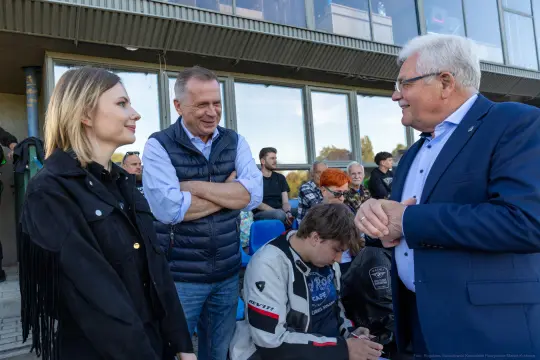 Spotkanie trzech osób, w tym mężczyzny w kamizelce i kobiety z długimi włosami. W tle widoczni inni ludzie.