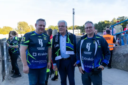 Trzech mężczyzn w strojach sportowych, z uśmiechami, na tle stadionu.