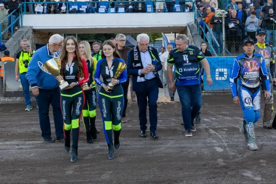 Kobiety niosące trofea, w tle tłum kibiców i zawodnicy na torze.
