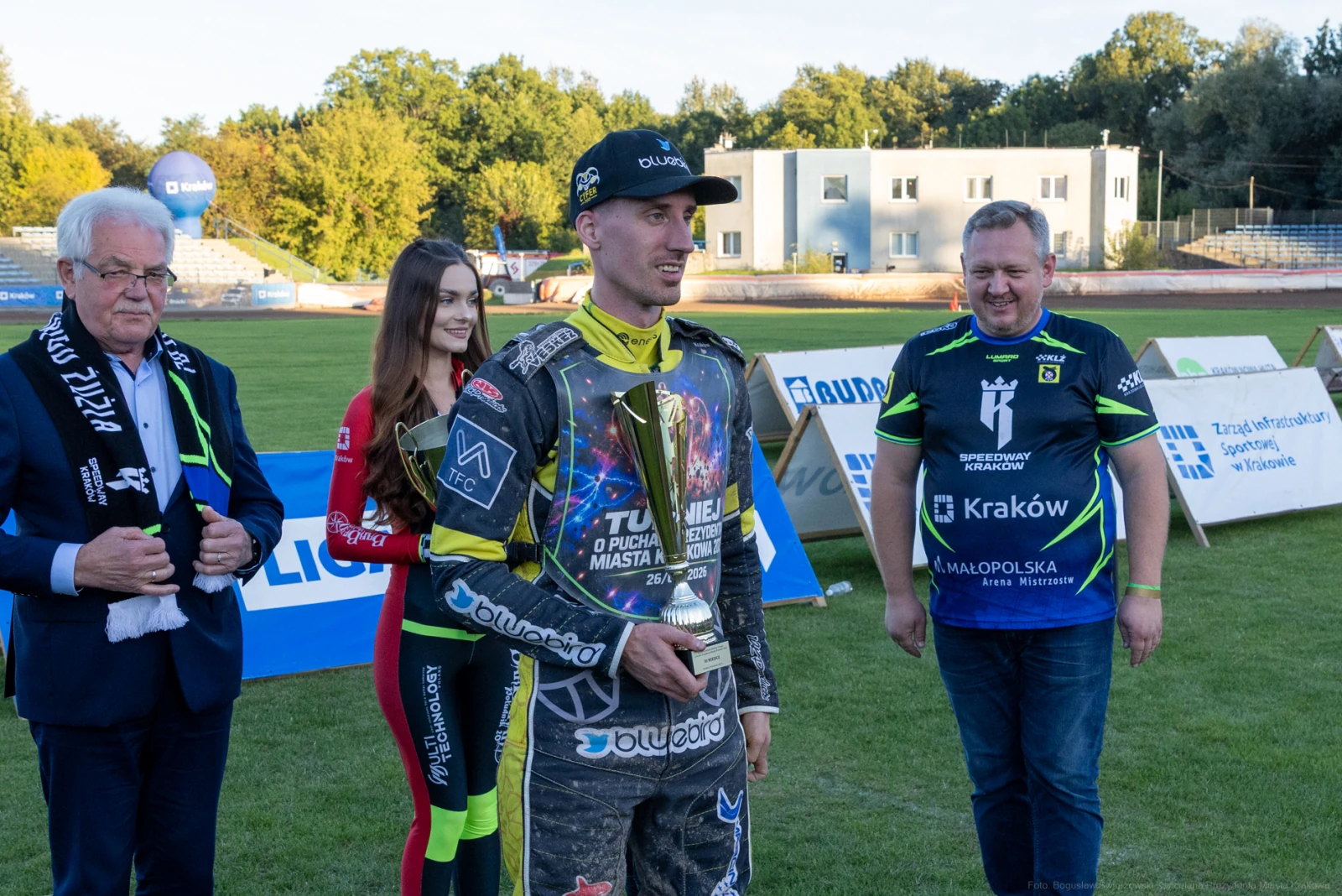 Zawodnik z pucharem, obok osoby w garniturze i kobiety w kombinezonie. Stadion w tle.  Autor: Bogusław Świerzowski
