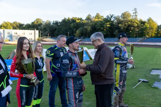 Zawodnicy speedwaya odbierają trofea po zawodach, w tle kibice i organizatorzy.
