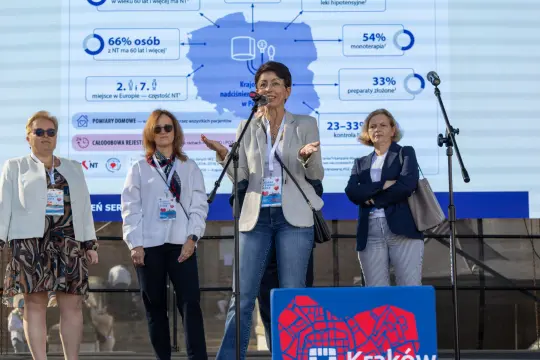 Kobieta przemawia na tle planszy z danymi statystycznymi, obok stoją trzy inne kobiety.