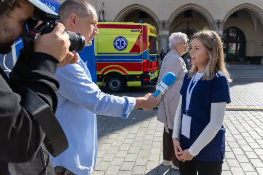 Dziennikarz przeprowadza wywiad z osobą w niebieskiej koszulce, w tle karetka i starsza kobieta.