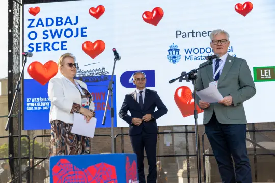 Kobieta i mężczyzna przemawiają na scenie podczas wydarzenia promującego zdrowie. W tle serca i logotypy partnerów.