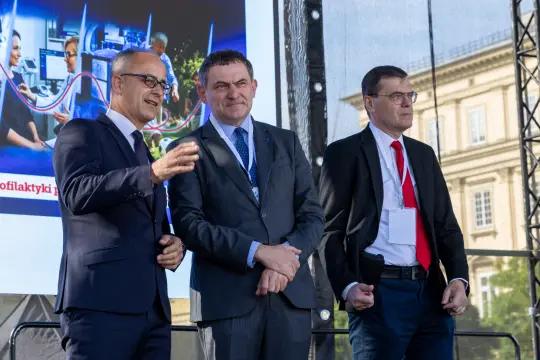 Trzech mężczyzn w garniturach podczas wydarzenia, rozmawiających na tle prezentacji.