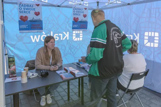 Kobieta przy stoisku informacyjnym, rozmowa z mężczyzną, ulotki o zdrowiu serca.