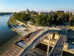 Nowoczesny most nad rzeką, z widokiem na zieleń i zabudowę miejską.