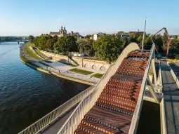 Nowoczesny most nad rzeką z widokiem na zieleń i zabudowę miejską.