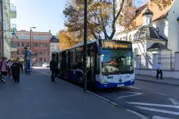 Autobus na przystanku w mieście, obok budynki i drzewa w jesiennych kolorach.
