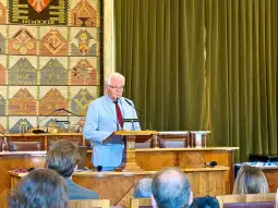 Mężczyzna w jasnym garniturze przemawia z mównicy w sali obrad.