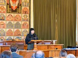 Kobieta przemawia z mównicy w sali z dekoracyjnymi tkaninami na ścianach.