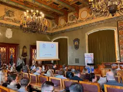 Sala konferencyjna z uczestnikami, ekranem prezentacyjnym i dekoracjami na ścianach.