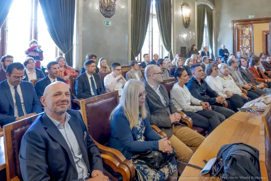 Grupa ludzi siedzących w sali, uczestniczących w wydarzeniu formalnym.