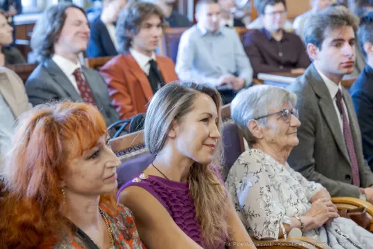 Grupa osób siedzących w sali, uśmiechnięte twarze, różnorodne stroje.