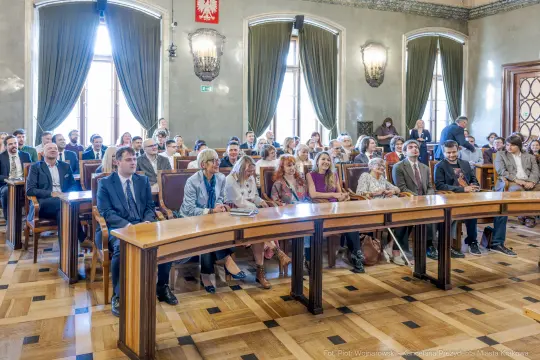 Grupa ludzi siedzących w sali, uczestniczących w wydarzeniu formalnym.