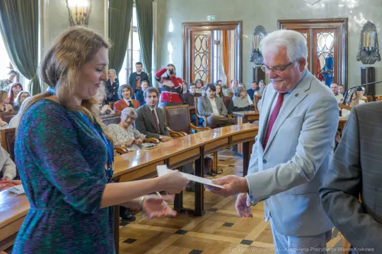 Wręczenie dyplomu przez mężczyznę kobiecie w sali obrad z zebranymi gośćmi.