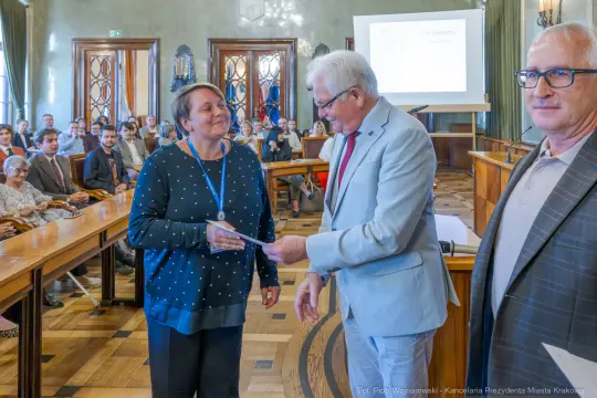 Wręczenie nagrody w sali obrad, uśmiechnięte osoby, dyplom w rękach.