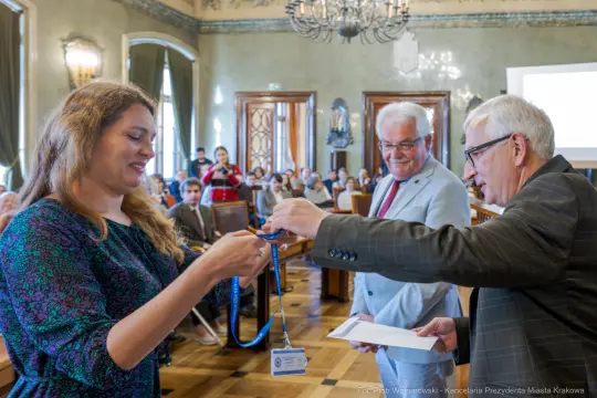 Wręczenie nagrody przez dwóch mężczyzn kobiecie w sali z publicznością.