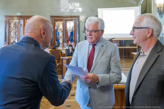 Wręczenie dokumentów w sali konferencyjnej, trzech mężczyzn w formalnych strojach.