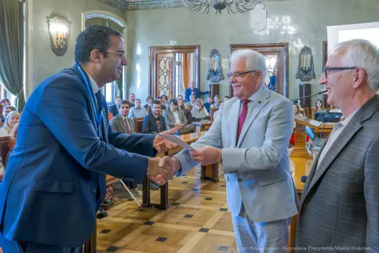 Wręczenie dyplomu w sali obrad z uśmiechniętymi uczestnikami.
