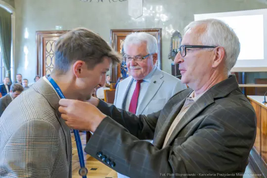 Mężczyzna odbiera medal od drugiego mężczyzny w obecności trzeciego.