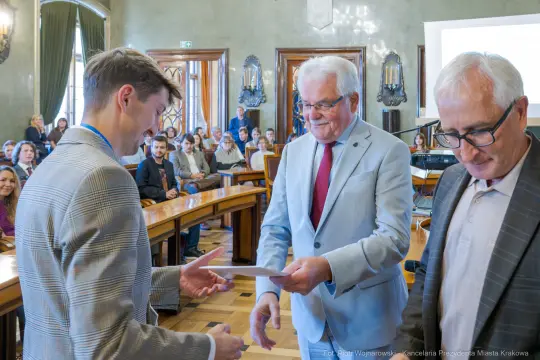 Wręczenie nagrody w sali obrad, trzech mężczyzn w formalnych strojach, publiczność w tle.