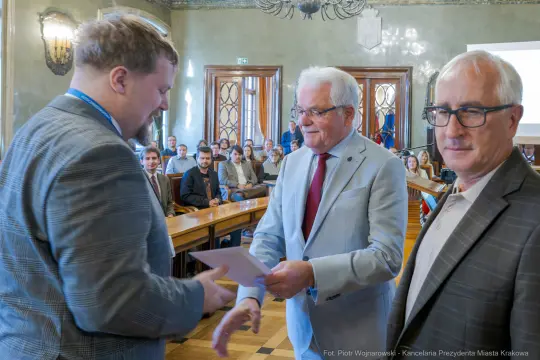 Wręczenie dyplomu w sali obrad z udziałem trzech mężczyzn.