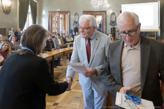 Wręczenie nagród podczas uroczystości w sali obrad, uściski dłoni i uśmiechy uczestników.
