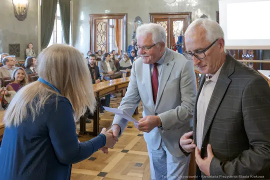 Wręczenie nagrody podczas uroczystości w sali obrad z uczestnikami w tle.