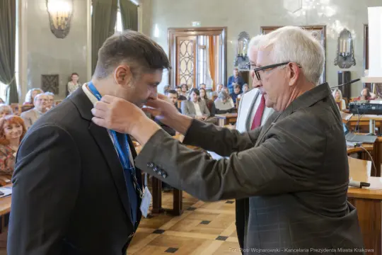 Mężczyzna odbiera medal w sali obrad, w tle zgromadzeni goście.