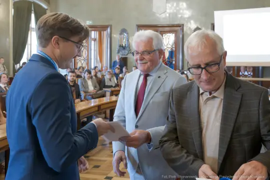 Wręczenie nagród w sali obrad, trzech mężczyzn w formalnych strojach.