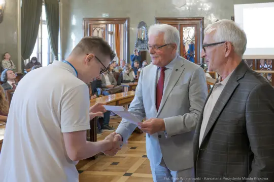 Wręczenie dyplomu młodemu laureatowi przez dwóch mężczyzn w sali obrad.