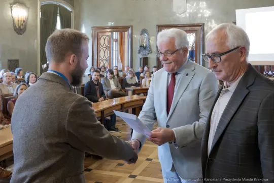 Mężczyzna odbierający dyplom w sali obrad z uśmiechniętymi mężczyznami w garniturach.