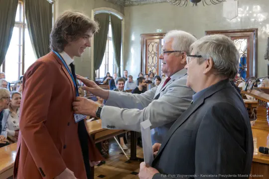 Wręczenie nagrody młodemu mężczyźnie w sali obrad z uczestnikami w tle.
