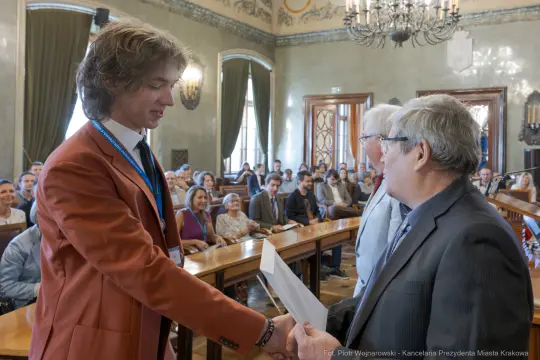 Młody mężczyzna odbiera dyplom w sali obrad, w tle zgromadzeni goście.