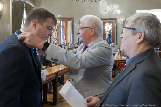 Ceremonia wręczenia odznaczenia w sali obrad z udziałem trzech mężczyzn.