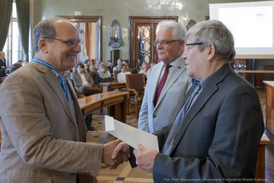Mężczyźni w garniturach wymieniają uściski dłoni i dokumenty w sali konferencyjnej.