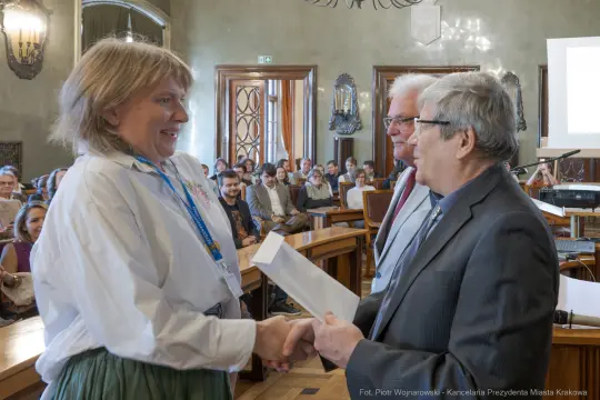 Wręczenie nagrody podczas uroczystości w sali obrad z uczestnikami w tle.