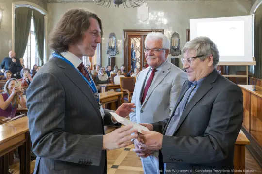 Wręczenie nagrody w sali obrad, uśmiechnięci mężczyźni w formalnych strojach.