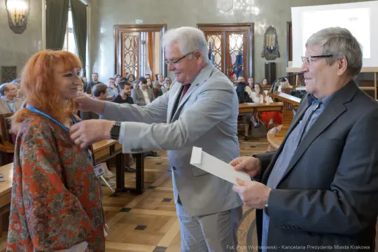 Wręczenie nagrody kobiecie przez dwóch mężczyzn w sali z publicznością.