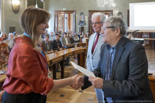 Wręczenie dyplomu w sali obrad, uściski dłoni i uśmiechy uczestników.