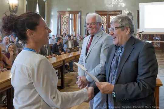 Wręczenie nagrody w sali obrad, uśmiechnięte twarze, gratulacje.