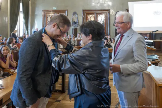 Uroczystość wręczenia odznaczenia w sali obrad z uśmiechniętymi uczestnikami.