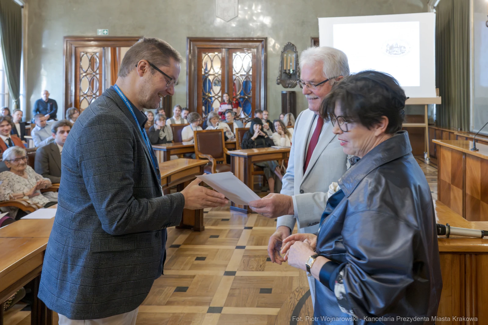 Wręczenie nagrody w sali obrad, trzech osób przy stole, dokument w rękach mężczyzny.  Autor: Piotr Wojnarowski